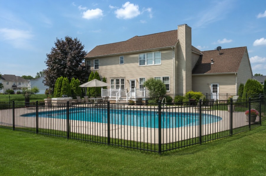Black aluminum picket fence enclosing a backyard swimming pool at a suburban New Jersey home on a sunny day, with green lawn, landscaped yard, and natural daylight creating a clean, photorealistic outdoor setting.