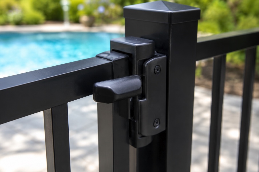 Close-up of a self-latching pool gate latch mounted on a black aluminum fence, with blurred blue swimming pool water in the background, captured in bright natural daylight with shallow depth of field and photorealistic detail.
