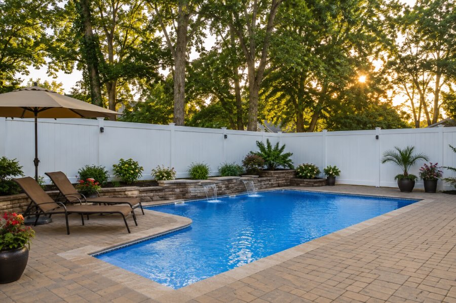 Residential backyard swimming pool enclosed by a white vinyl privacy fence, with mature trees rising above the fence line, landscaped patio area, and warm late afternoon sunlight creating a calm, photorealistic outdoor setting.